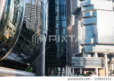 Curved reflective glass building exterior facade stands directly alongside rigid octagonal geometric skyscraper creating strong architectural contrast inside dense modern corporate business district Curved reflective glass building exterior facade stands directly alongside rigid octagonal geometric skyscraper creating strong architectural contrast inside dense modern corporate business district 139661051