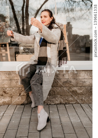 Woman winter fashion city stylish lady in checkered coat and scarf sitting outdoors during a peaceful morning in town Woman winter fashion city stylish lady in checkered coat and scarf sitting outdoors during a peaceful morning in town 139661338