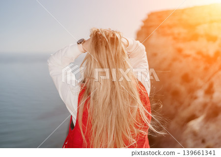 Woman sunset cliff standing on the edge overlooking sea holding long blonde hair expressing freedom during golden hour Woman sunset cliff standing on the edge overlooking sea holding long blonde hair expressing freedom during golden hour 139661341