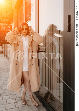 Fashion model street. Stylish woman in beige trench coat and sunglasses posing by glass building with warm sunlight Fashion model street. Stylish woman in beige trench coat and sunglasses posing by glass building with warm sunlight 139661342