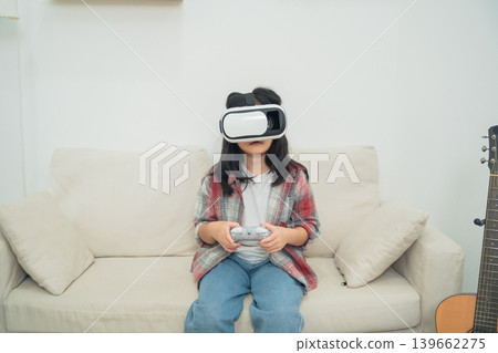 Young child wearing virtual reality headset while sitting on a couch with a game controller in a cozy living room environment, engaging in immersive gaming experience Young child wearing virtual reality headset while sitting on a couch with a game controller in a cozy living room environment, engaging in immersive gaming experience 139662275