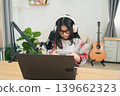Young girl wearing headphones and glasses, engaged in gaming with a controller, sitting at a desk with a laptop and microphone in a cozy room setting 139662323