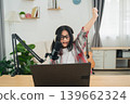 Young girl with glasses celebrating success while using a laptop in a cozy home office setting with a microphone and plants around her 139662324
