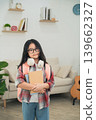 Young girl with glasses and backpack standing in a cozy living room, holding a notebook, ready for school, wearing a plaid shirt, bright and cheerful atmosphere 139662327