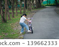Young girl learning to ride a bicycle in a serene park environment, surrounded by trees and greenery, showcasing childhood innocence and adventure 139662333