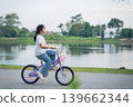 Happy girl riding a purple bicycle near a serene lake with lush greenery, enjoying a warm sunny day and embracing the joy of childhood and adventure 139662344