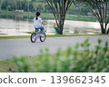 Child Riding Bicycle by Calm Lake in Park Surrounded by Lush Trees, Enjoying Outdoor Activity during Sunny Day with Clear Sky and Peaceful Atmosphere 139662345