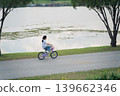 Young girl riding a bicycle along a scenic path beside a tranquil lake, surrounded by lush greenery and trees in a peaceful outdoor setting 139662346