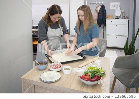 Cheerful daughter and mother cooking italian pizza together in cozy domestic kitchen. Cheerful daughter and mother cooking italian pizza together in cozy domestic kitchen. 139663596