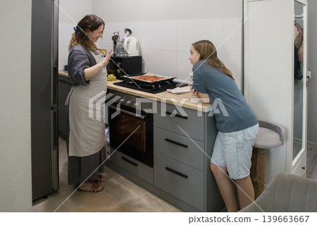 Positive mother and daughter cooking italian pizza together in cozy home kitchen. Positive mother and daughter cooking italian pizza together in cozy home kitchen. 139663667