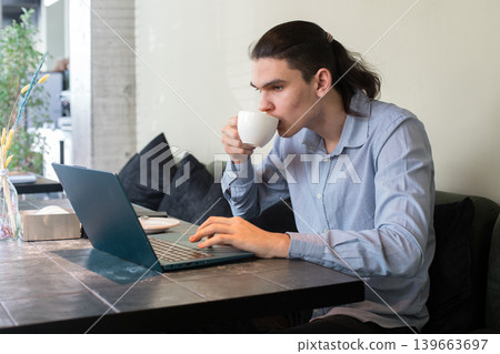 A young man sits at a cafe table focused on his laptop while sipping coffee. 139663697
