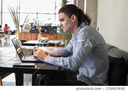 A young man sits at a cafe table focused on his laptop while sipping coffee. A young man sits at a cafe table focused on his laptop while sipping coffee. 139663698