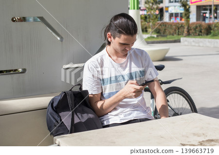 A young man sits on a bench at a bus stop in an urban park. He is focused on his phone A young man sits on a bench at a bus stop in an urban park. He is focused on his phone 139663719