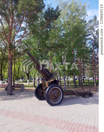 Historic Soviet-era howitzer stands as a monument in a sunny city park. Surrounded by lush green trees and paved walkways, this military relic serves as a peaceful memorial of past bravery 139664514