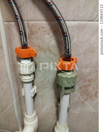 Flexible braided metal hoses are connected to white plastic pipes using orange butterfly-style wing nuts. This plumbing setup is mounted against a tiled wall, showing a typical home water installation 139664515