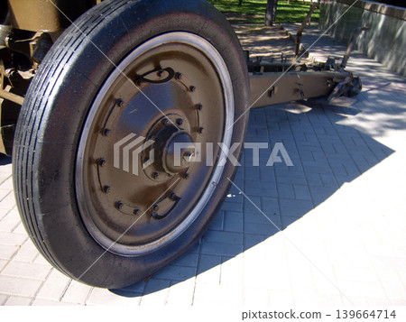 Close-up view of a massive rubber tire and metal rim belonging to a vintage military artillery piece. The weathered cannon stands as a park monument, casting deep shadows on the paved ground Close-up view of a massive rubber tire and metal rim belonging to a vintage military artillery piece. The weathered cannon stands as a park monument, casting deep shadows on the paved ground 139664714
