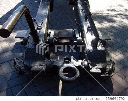 Close-up view shows the heavy iron hitch and trails of a vintage Soviet artillery gun. Positioned on a paved walkway, the dark metal structure reflects bright sunlight and deep shadows Close-up view shows the heavy iron hitch and trails of a vintage Soviet artillery gun. Positioned on a paved walkway, the dark metal structure reflects bright sunlight and deep shadows 139664792