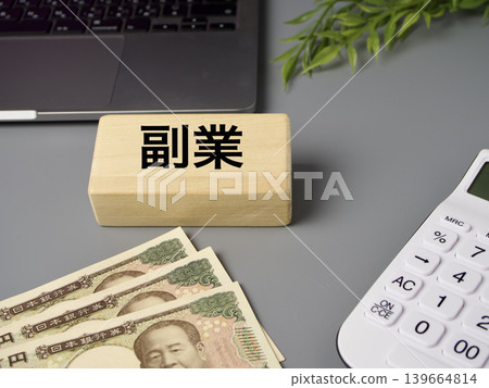 A business image featuring wooden blocks, a calculator, and banknotes, representing a side hustle, against a gray background. 139664814