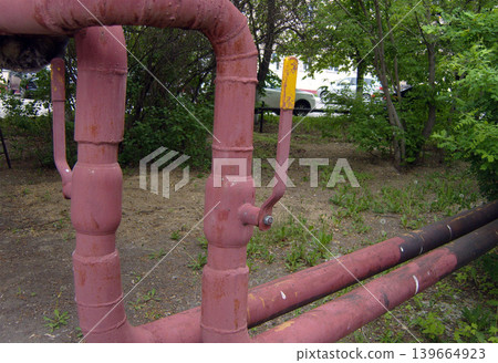 Old red metal pipes with yellow-tipped valve handle stand in grassy urban area. Weathered pipes, showing signs of rust and age, are part of local heating or gas distribution system. Old red metal pipes with yellow-tipped valve handle stand in grassy urban area. Weathered pipes, showing signs of rust and age, are part of local heating or gas distribution system. 139664923