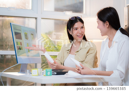 Two asian woman discussing cosmetic tube packaging sitting at modern home office. 139665385