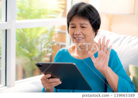 Happy senior asian woman sitting on sofa video call on tablet in living room at home. Happy senior asian woman sitting on sofa video call on tablet in living room at home. 139665389