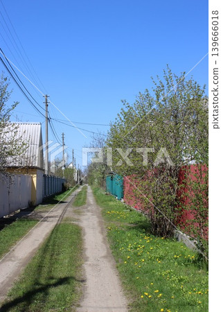 Dirt path lined with green grass and flowering dandelions, flanked by colorful fences and trees under a clear blue sky in a rural neighborhood Dirt path lined with green grass and flowering dandelions, flanked by colorful fences and trees under a clear blue sky in a rural neighborhood 139666018