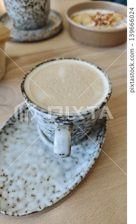 Coffee cup with speckled ceramic design resting on a matching saucer, placed on a wooden table with a bowl of sliced bananas in the background Coffee cup with speckled ceramic design resting on a matching saucer, placed on a wooden table with a bowl of sliced bananas in the background 139666024