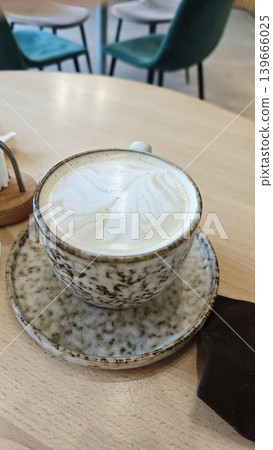 Latte art in a coffee cup on a saucer sits on a wooden table in a contemporary cafe, with blurred chairs and ambient lighting visible in the background Latte art in a coffee cup on a saucer sits on a wooden table in a contemporary cafe, with blurred chairs and ambient lighting visible in the background 139666025