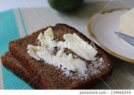 Dark rye bread slices topped with butter on a textured tablecloth, accompanied by a plate of butter and a green apple in the background Dark rye bread slices topped with butter on a textured tablecloth, accompanied by a plate of butter and a green apple in the background 139666059