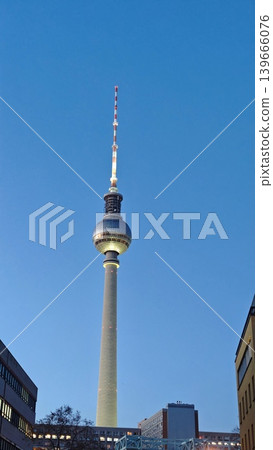 Berlin TV Tower illuminated at dusk, showcasing its iconic structure against a clear blue sky with surrounding buildings partially visible in the foreground 139666076