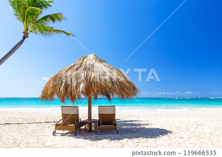 Sunny tropical beach with lounge chairs and palm umbrella in Punta Cana, Dominican republic. Perfect vacation destination with white sand, turquoise ocean and clear blue sky. Sunny tropical beach with lounge chairs and palm umbrella in Punta Cana, Dominican republic. Perfect vacation destination with white sand, turquoise ocean and clear blue sky. 139666535
