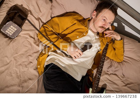 Man lies on bed testing voice ideas using his phone and guitar for demo Man lies on bed testing voice ideas using his phone and guitar for demo 139667238