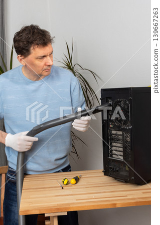 A 53-year-old man cleans dust from a computer system unit with a vacuum cleaner in a tidy room. He carefully removes dirt to keep the device running well A 53-year-old man cleans dust from a computer system unit with a vacuum cleaner in a tidy room. He carefully removes dirt to keep the device running well 139667263