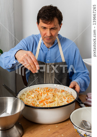 In a kitchen, a 53-year-old man is adding salt to a mixture of cabbage and carrots in a large bowl. He focuses on the fermentation process that supports gut health In a kitchen, a 53-year-old man is adding salt to a mixture of cabbage and carrots in a large bowl. He focuses on the fermentation process that supports gut health 139667265