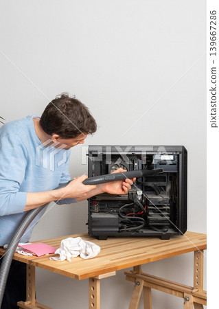 A 53-year-old man uses a vacuum to clean dust from a computer system unit on a wooden table in a home office. The setting is tidy and organized 139667286