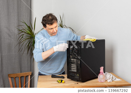 A 53-year-old man uses a cloth to clean dust from a computer system unit. He works at a wooden table in a room with plants nearby. Tools are set out for the task A 53-year-old man uses a cloth to clean dust from a computer system unit. He works at a wooden table in a room with plants nearby. Tools are set out for the task 139667292