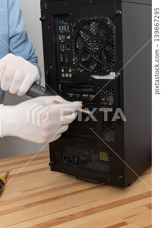 A 53-year-old man removes dust from a computer system unit using a brush while wearing gloves. He is focused on maintaining the computer in a room with plants A 53-year-old man removes dust from a computer system unit using a brush while wearing gloves. He is focused on maintaining the computer in a room with plants 139667295