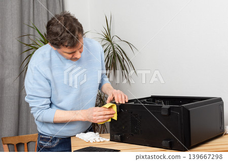 A 53-year-old man uses a cloth to clean dust from a computer system unit. He works at a wooden table in a room with plants nearby. Tools are set out for the task A 53-year-old man uses a cloth to clean dust from a computer system unit. He works at a wooden table in a room with plants nearby. Tools are set out for the task 139667298