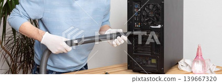 A 53-year-old man cleans dust from a computer system unit with a vacuum cleaner in a tidy room. He carefully removes dirt to keep the device running well, banner 139667320