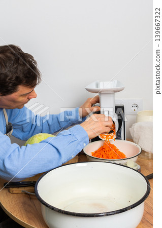 A 53-year-old man is in his kitchen preparing cabbage and carrots for fermentation. He uses a machine to shred the vegetables before placing them in a bowl A 53-year-old man is in his kitchen preparing cabbage and carrots for fermentation. He uses a machine to shred the vegetables before placing them in a bowl 139667322