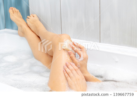 Close up of female legs in bubble bath, washing skin with natural soap bar. Woman enjoying a relaxing bath, creating gentle bubbles in the warm water, focusing on personal hygiene and skin care Close up of female legs in bubble bath, washing skin with natural soap bar. Woman enjoying a relaxing bath, creating gentle bubbles in the warm water, focusing on personal hygiene and skin care 139667759