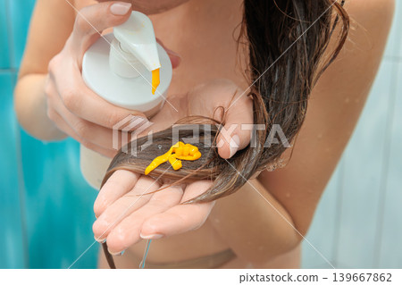 Close up woman standing in a shower, dispensing yellow hair conditioner from a pump bottle onto a section of her long, wet brown hair, preparing for a revitalizing hair care treatment Close up woman standing in a shower, dispensing yellow hair conditioner from a pump bottle onto a section of her long, wet brown hair, preparing for a revitalizing hair care treatment 139667862