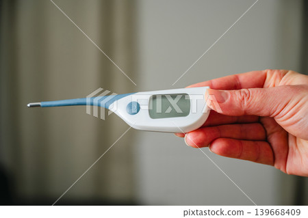 Electronic thermometer in female hands. Woman holding a digital thermometer. Flu and cold concept. Seasonal viruses Electronic thermometer in female hands. Woman holding a digital thermometer. Flu and cold concept. Seasonal viruses 139668409