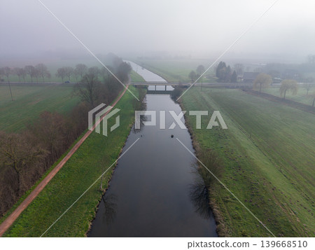 High-angle aerial view of a calm river canal flowing through a misty, foggy agricultural landscape in winter or early spring. A rural road and bridge cross the water, surrounded by bare trees. High-angle aerial view of a calm river canal flowing through a misty, foggy agricultural landscape in winter or early spring. A rural road and bridge cross the water, surrounded by bare trees. 139668510