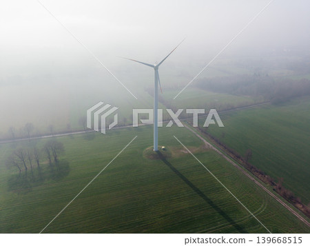 Aerial view of a solitary wind turbine standing in vast green agricultural fields on a foggy, misty morning. Clean energy technology in a rural environment. Aerial view of a solitary wind turbine standing in vast green agricultural fields on a foggy, misty morning. Clean energy technology in a rural environment. 139668515
