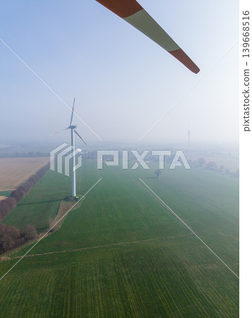 Aerial view looking through the rotating blade of a wind turbine, showing a single turbine standing in a misty, agricultural landscape. Sustainable green energy concept in fog. Aerial view looking through the rotating blade of a wind turbine, showing a single turbine standing in a misty, agricultural landscape. Sustainable green energy concept in fog. 139668516