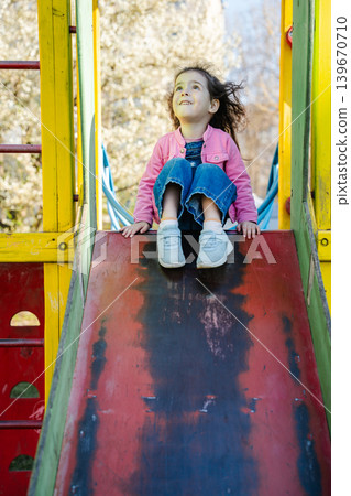 Caucasian girl sitting on playground slide in sunny residential park area. Childhood and outdoor lifestyle Caucasian girl sitting on playground slide in sunny residential park area. Childhood and outdoor lifestyle 139670710