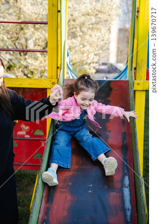 Caucasian girl sliding down playground slide with mother support in sunny park area. Childhood and family lifestyle Caucasian girl sliding down playground slide with mother support in sunny park area. Childhood and family lifestyle 139670717