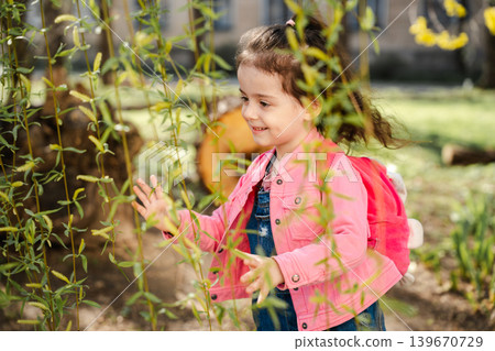 Caucasian girl with pink backpack touching willow tree branches in sunny park area. Childhood and spring lifestyle Caucasian girl with pink backpack touching willow tree branches in sunny park area. Childhood and spring lifestyle 139670729