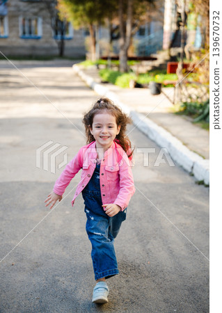 Caucasian girl in pink jacket running on residential street in sunny neighborhood area. Childhood and active lifestyle Caucasian girl in pink jacket running on residential street in sunny neighborhood area. Childhood and active lifestyle 139670732
